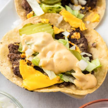 Two open-faced smash burger tacos topped with ground beef, melted cheese, chopped lettuce, onions, pickles, and a creamy burger sauce on corn tortillas.