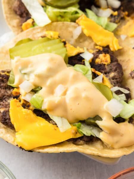 Two open-faced smash burger tacos topped with ground beef, melted cheese, chopped lettuce, onions, pickles, and a creamy burger sauce on corn tortillas.