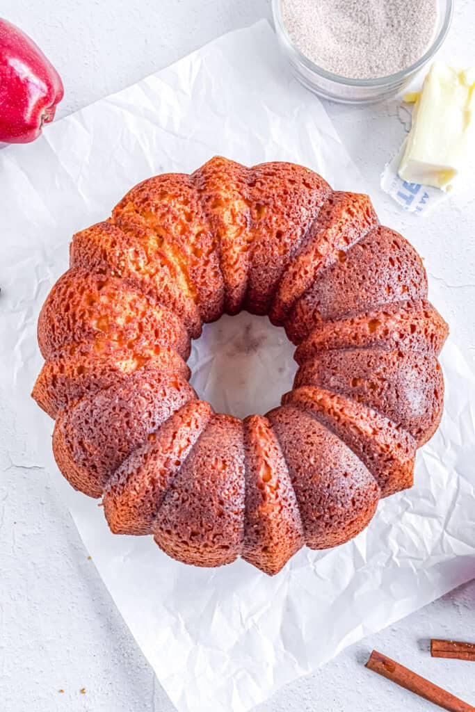 A golden-brown apple cider donut cake after being released from the pan with butter and cinnamon sugar nearby.