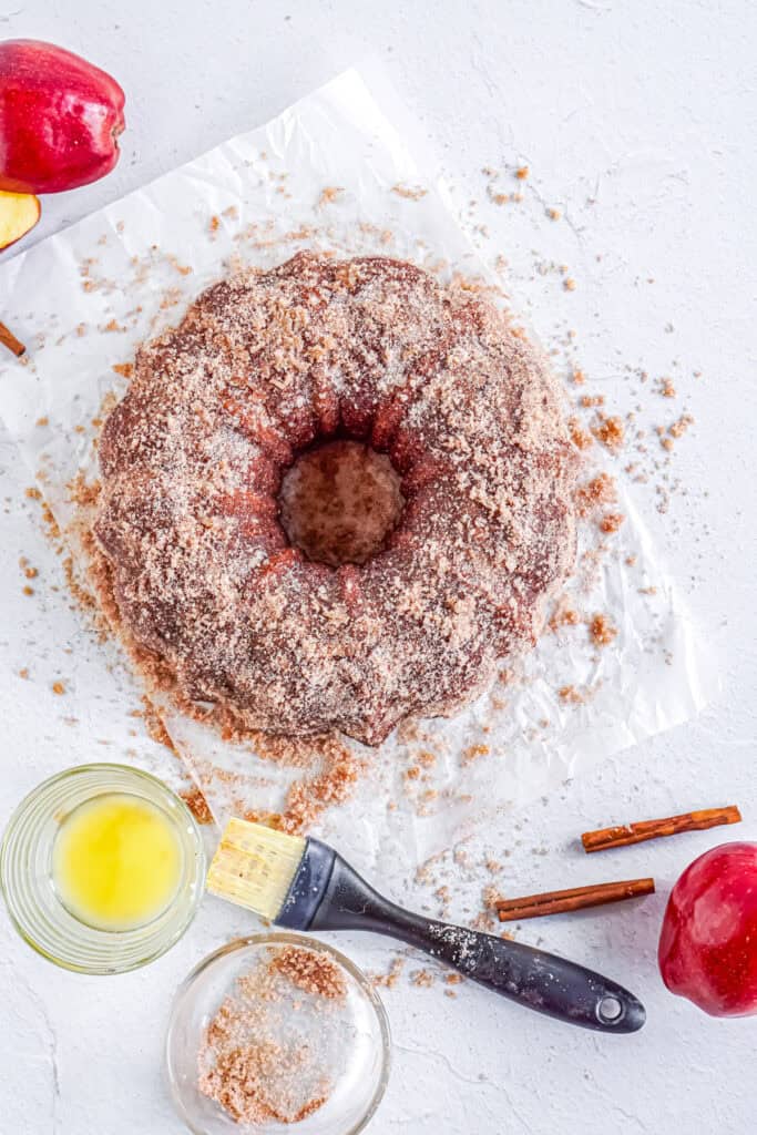 A round apple cider donut cake on parchment paper covered in melted butter and a thick layer of cinnamon sugar.