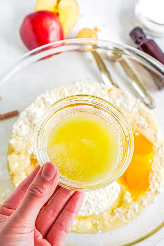 A hand holds a small bowl of melted butter above a mixing bowl with eggs, cake mix, and other ingredients for apple cider donut cake.