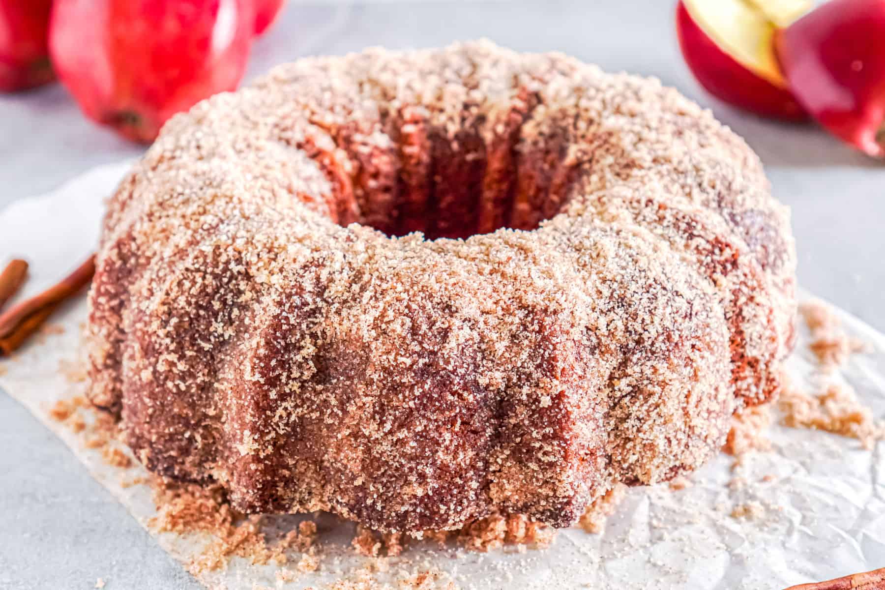 A round bundt cake coated with a layer of cinnamon sugar sits on parchment paper, with red apples and cinnamon sticks in the background.