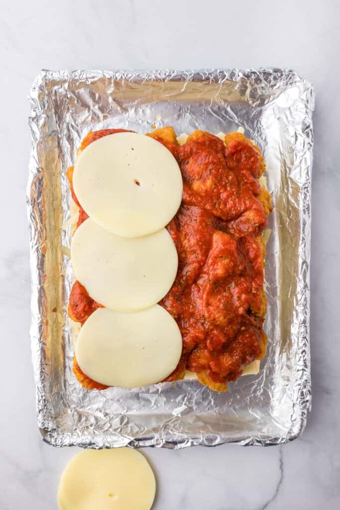 The bottom layer of a pack of Hawaiian rolls layered with three slices of provolone cheese being layered over cooked chicken nuggets, marinara and shredded mozzarella cheese. on a foil lined pan for chicken parmesan sliders.