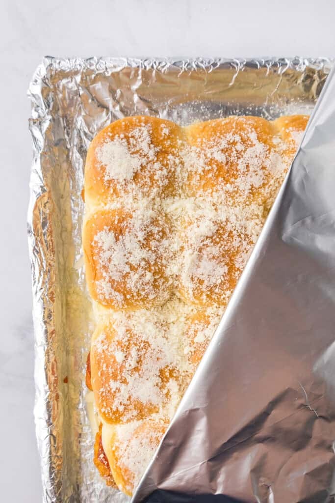 A tray of chicken parmesan sliders sprinkled with grated cheese being covered with aluminum foil over the pan.