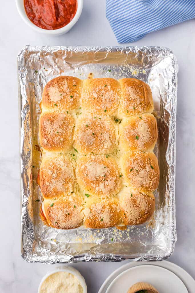 A tray of finished golden baked chicken parmesan slider topped with herbs and grated cheese on a foil lined pan.