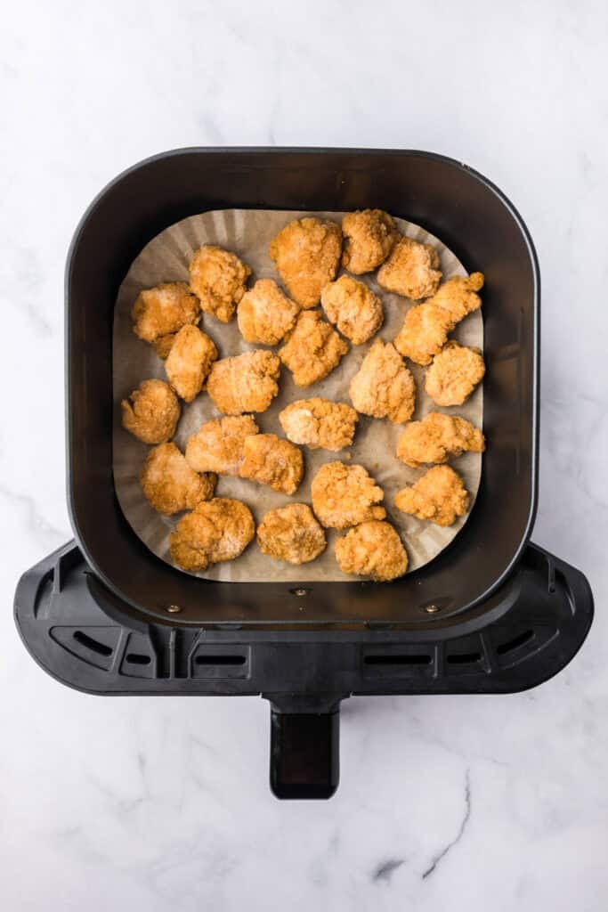 An air fryer basket lined with parchment paper with frozen chicken nuggets being cooked for chicken parmesan sliders.