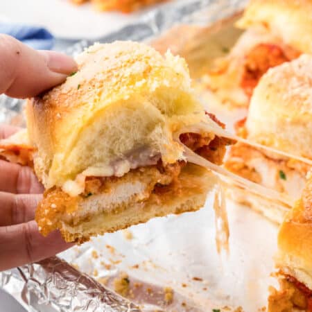 A hand holds a chicken parmesan slider with melted cheese stretching from the sandwichto more sliders on a foil-lined tray.