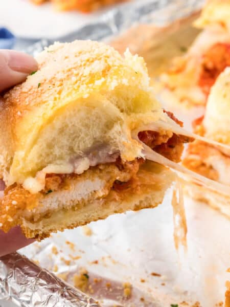 A hand holds a chicken parmesan slider with melted cheese stretching from the sandwichto more sliders on a foil-lined tray.