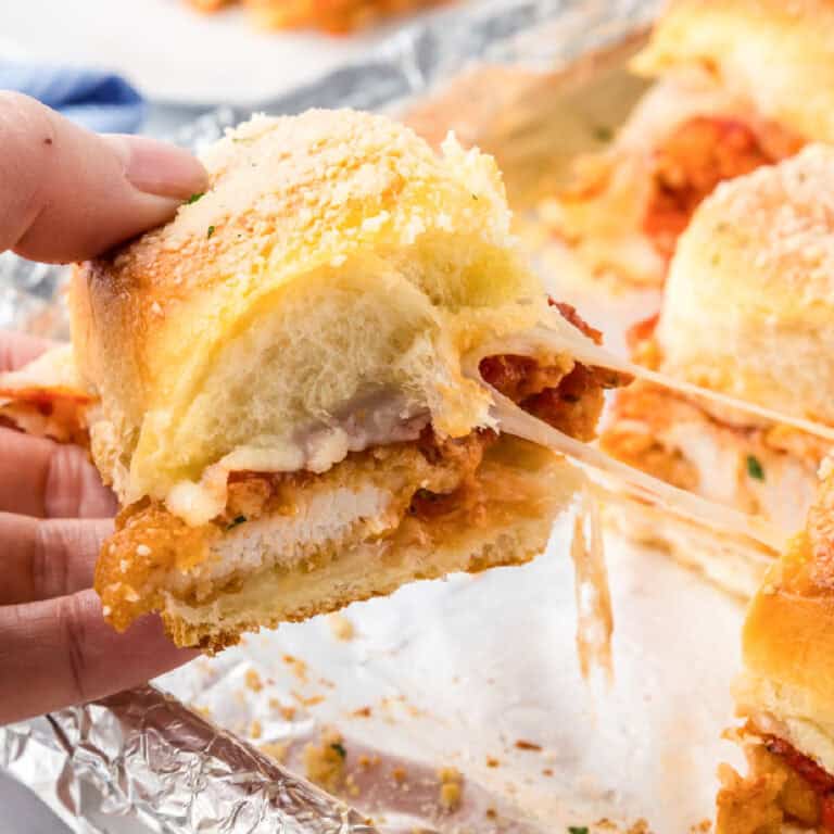 A hand holds a chicken parmesan slider with melted cheese stretching from the sandwichto more sliders on a foil-lined tray.