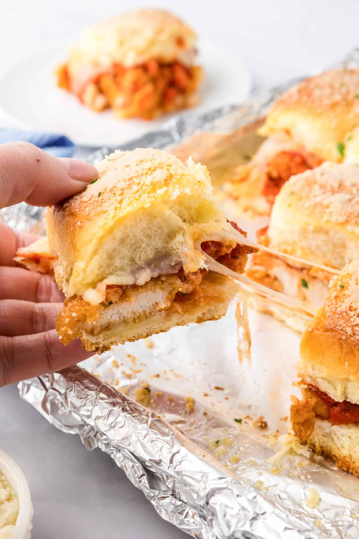 A hand holds a cheesy chicken parmesan slider sandwich pulled from a foil-lined tray with several more sandwiches.
