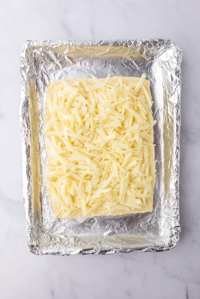 Shredded cheese covers the bottom layer of the Hawaiian rolls on a foil lined pan for chicken parmesan sliders.