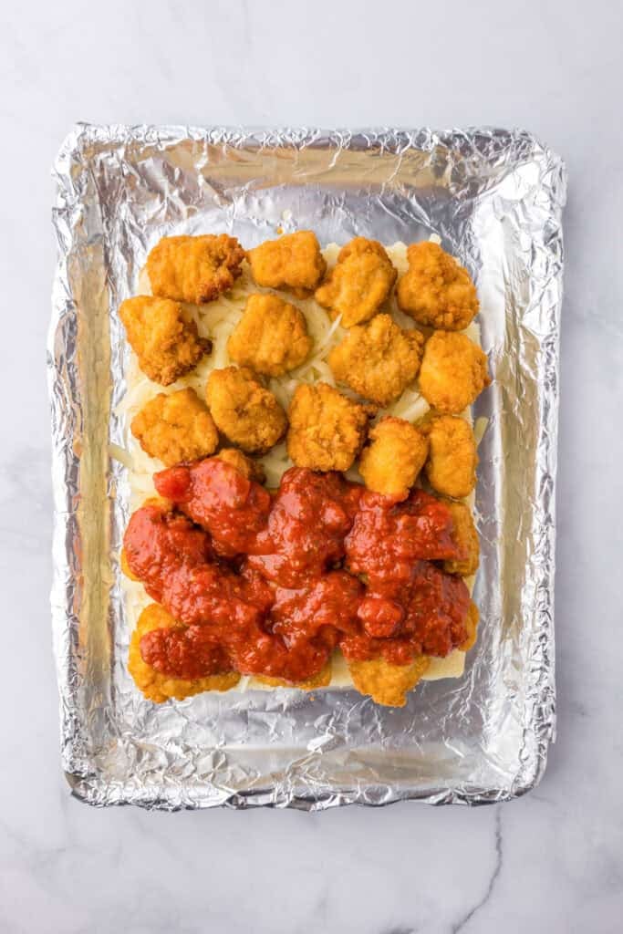The bottom layer of a pack of Hawaiian rolls layered with mozzarella cheese, cooked chicken nuggets with marinara being layered over the chicken nuggets on a foil lined pan for chicken parmesan sliders.