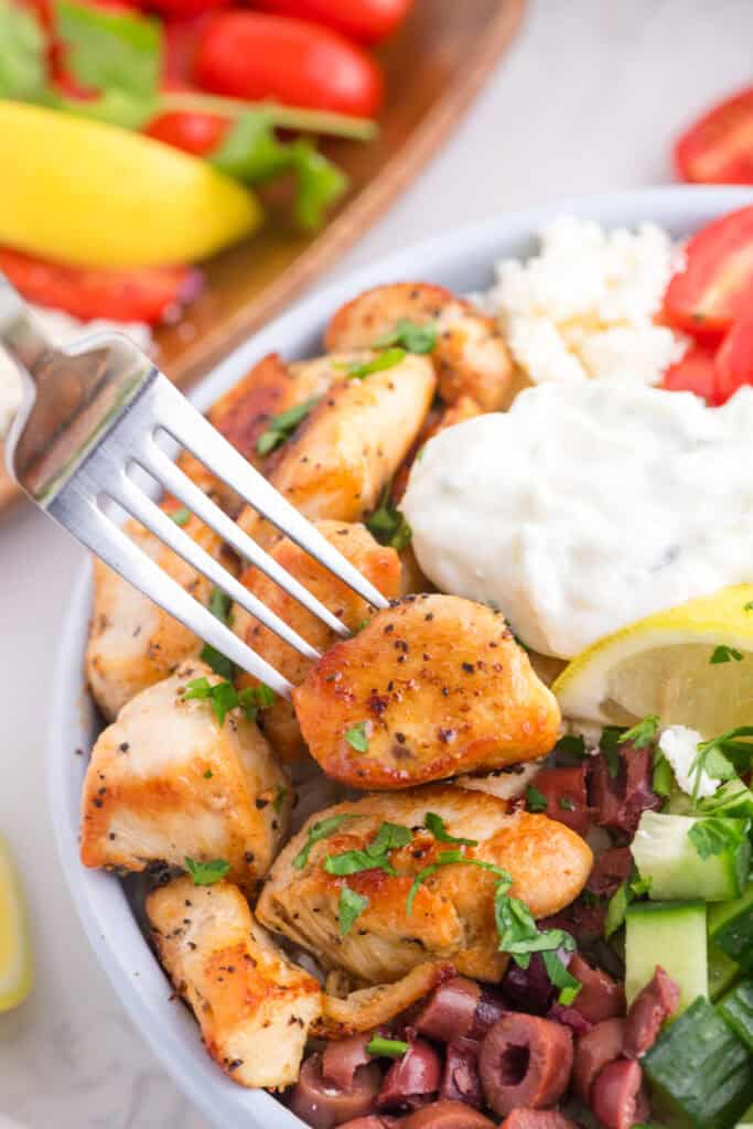 A fork close up holds a piece of seasoned chicken over a greek chicken bowl bowl with diced cucumbers, tomatoes, kalamata olives, feta cheese, rice and tzatziki sauce.