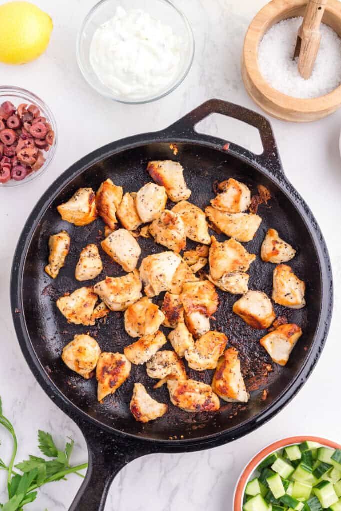 Chunks of cooked chicken breast in a black skillet, surrounded other toppings for greek chicken bowls.