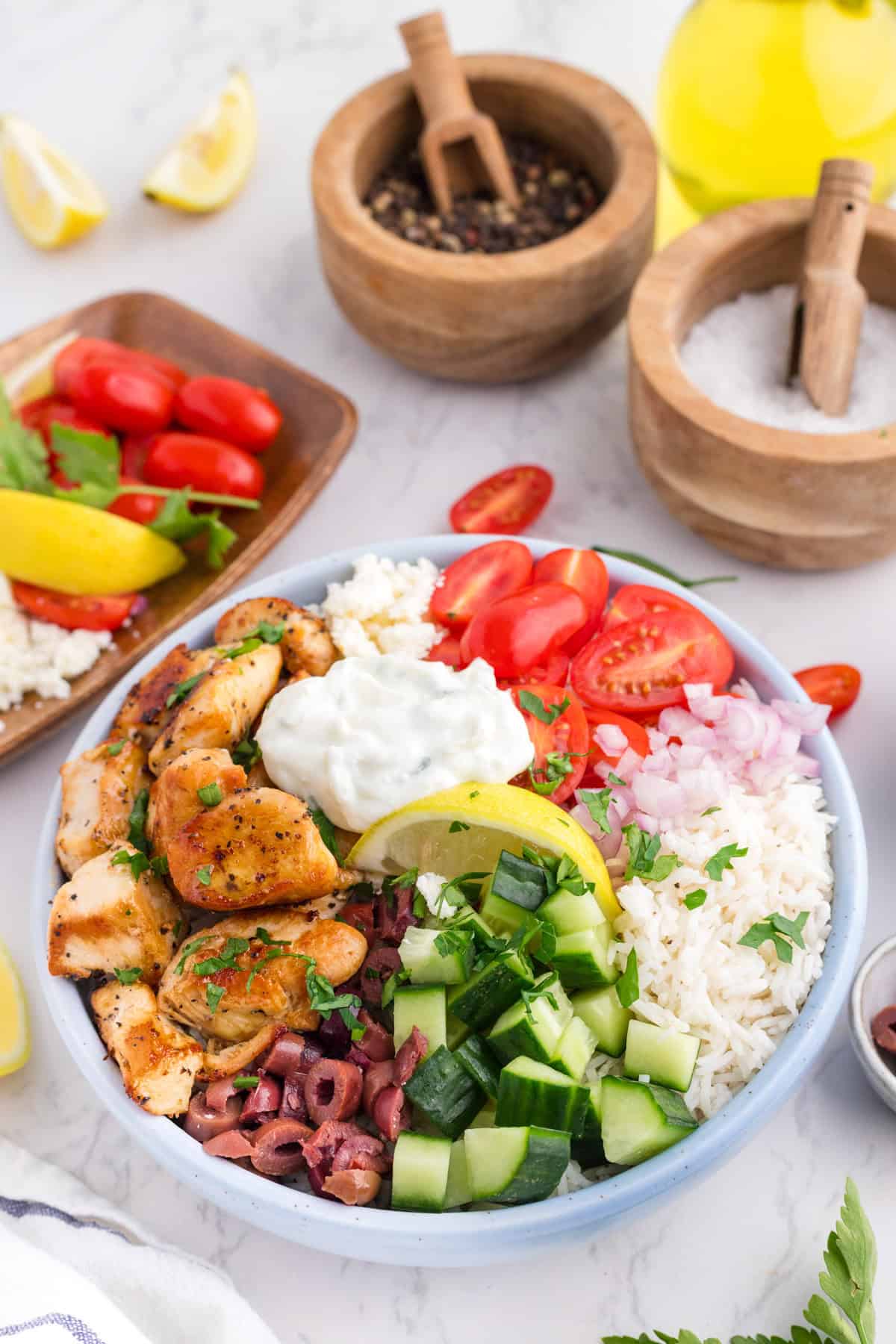 A greek chicken bowl after assembling with seasoned chicken, rice, cucumber, tomatoes, onions, olives, feta, and tzatziki on a countertop for dinner.