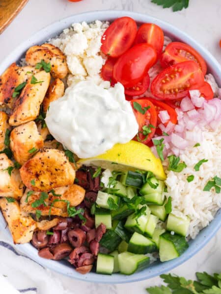 A Greek chicken bowl with seasoned chicken, cherry tomatoes, onions, rice, feta cheese, olives, diced cucumber, a lemon wedge, and a dollop of tzatziki sauce from overhead.