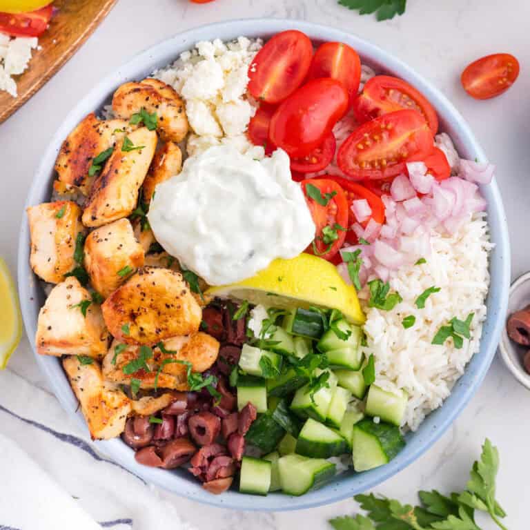 A Greek chicken bowl with seasoned chicken, cherry tomatoes, onions, rice, feta cheese, olives, diced cucumber, a lemon wedge, and a dollop of tzatziki sauce from overhead.