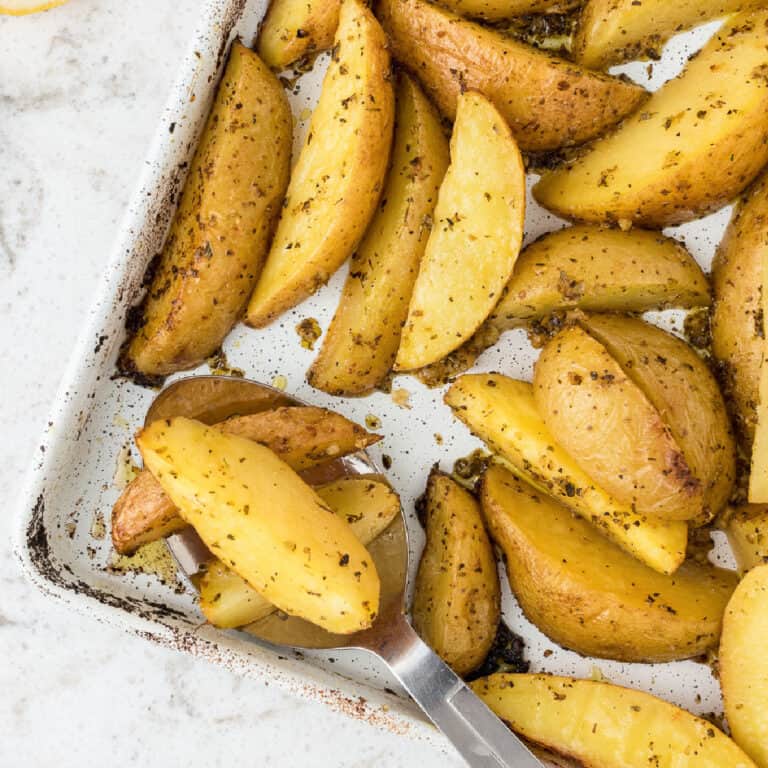 Baked potato wedges seasoned with herbs and a hint of lemon Greek potatoes flavor, served on a metal tray with a serving spatula lifting a portion.