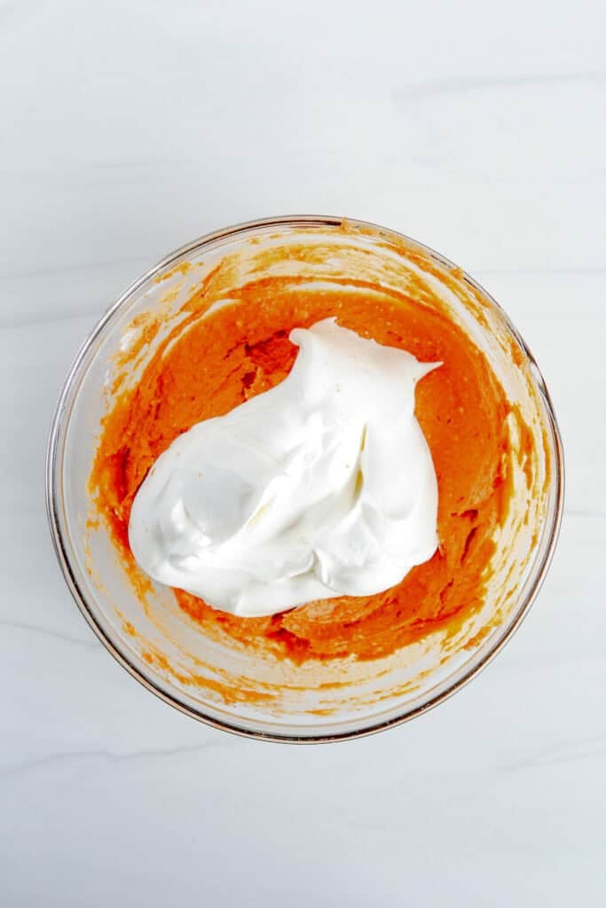 A glass bowl with orange pumpkin mixture and a large amount of Cool Whip being folded into the pumpkin pie dip.