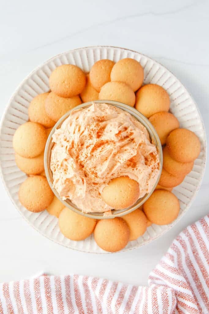 A bowl of creamy pumpkin pie dip dip sprinkled with pumpkin pie spice on a splatter surrounded with vanilla wafers for dipping.