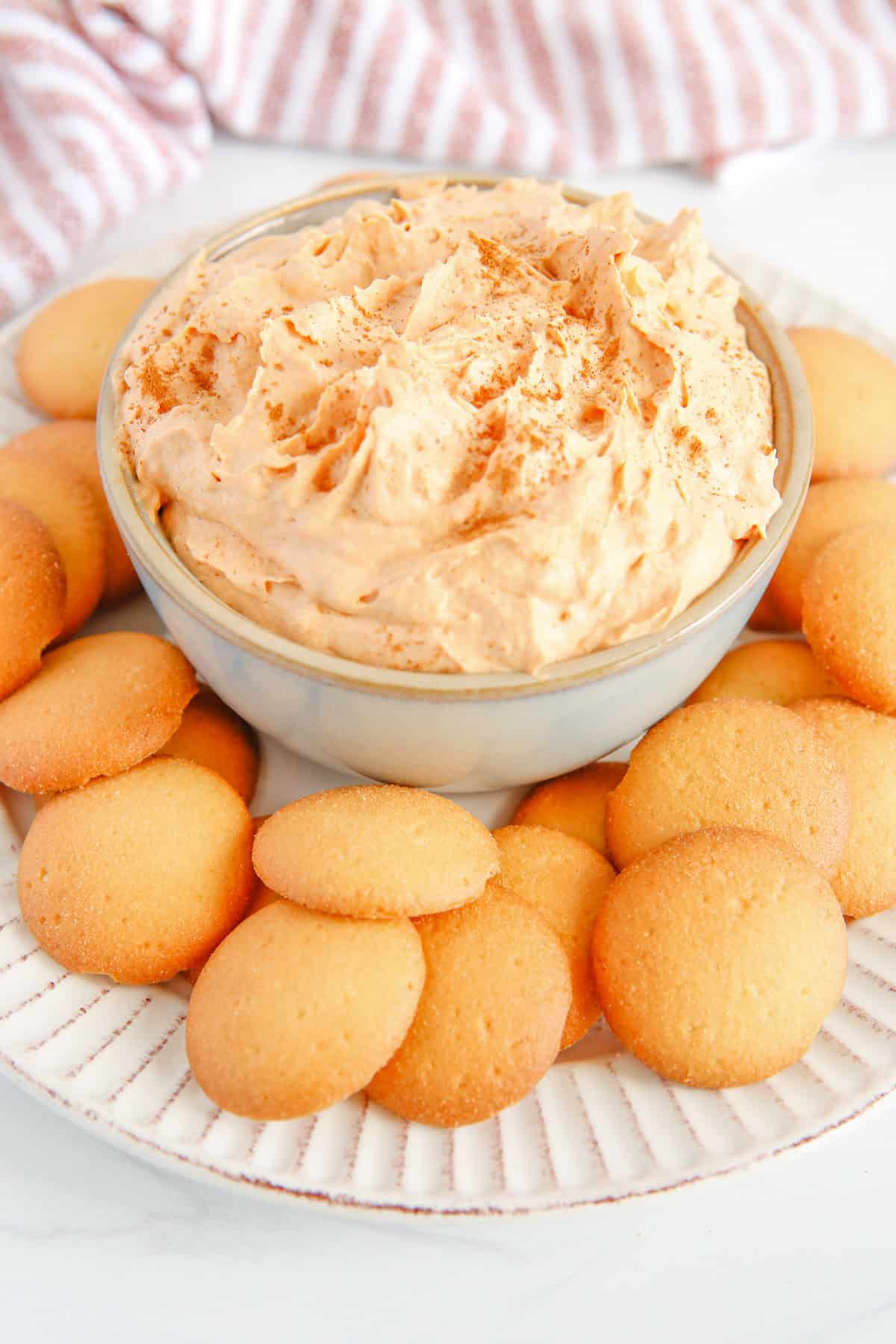 A finished dip sized bowl of creamy pumpkin pie dip topped with cinnamon, surrounded by round vanilla wafer cookies on a white plate.