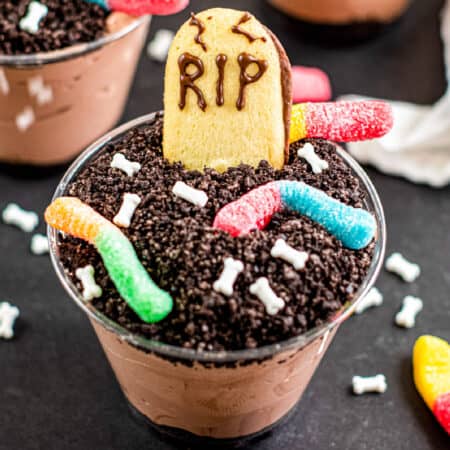 Square view of a chocolate graveyard dirt cup filled with chocolate pudding topped with crushed cookies, gummy worms, candy bones, and a cookie decorated as a gravestone with “RIP” written in chocolate.