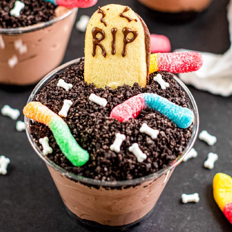 Square view of a chocolate graveyard dirt cup filled with chocolate pudding topped with crushed cookies, gummy worms, candy bones, and a cookie decorated as a gravestone with “RIP” written in chocolate.