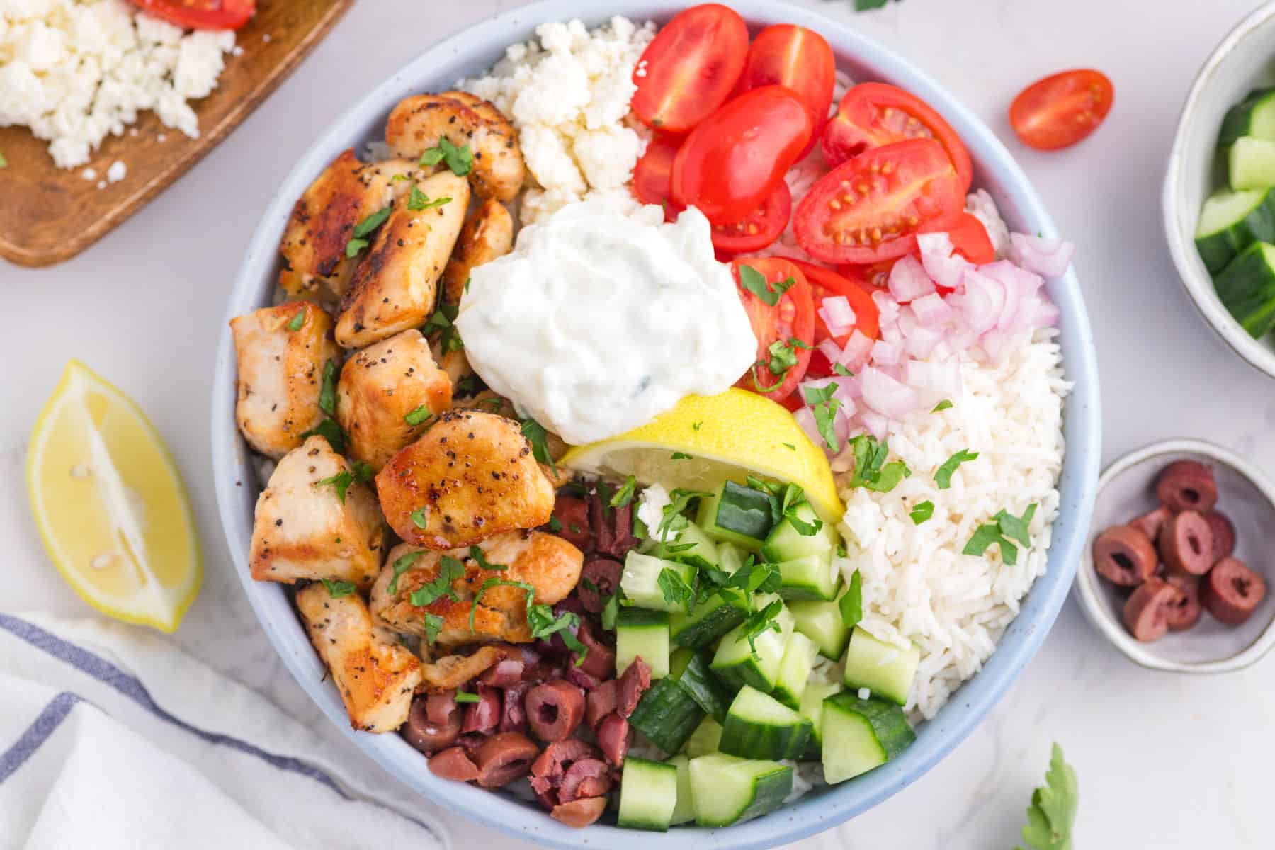 An overhead view of a greek chicken bowl with seasoned grilled chicken, rice, cherry tomatoes, cucumber, red onions, olives, feta cheese, a lemon wedge, and a dollop of tzatziki sauce on a counter.