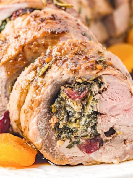 Sliced stuffed pork tenderloin with a spinach, cheese, and cranberry filling, served on a white plate with dried apricots and cranberries.