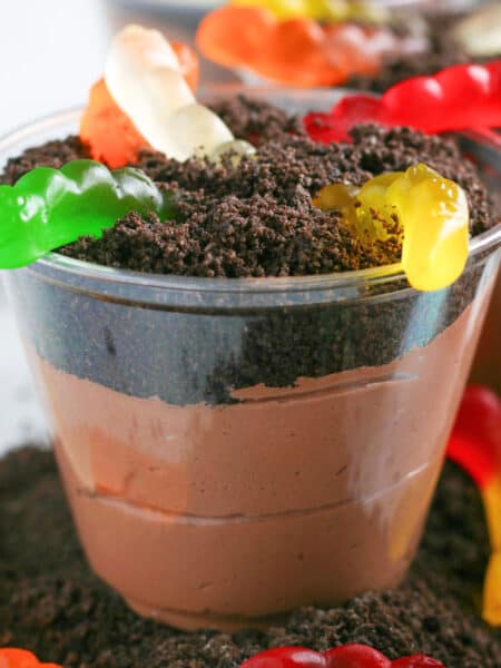 A plastic cup filled with chocolate pudding, topped with crushed cookies and several colorful gummy worms for chocolate dirt cups.