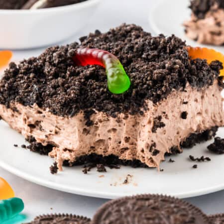 A square slice of chocolate Oreo dirt cake topped with crumbled cookies and a gummy worm, served on a white plate with extra gummy worms nearby.