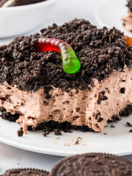 A square slice of chocolate Oreo dirt cake topped with crumbled cookies and a gummy worm, served on a white plate with extra gummy worms nearby.