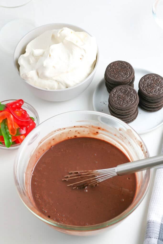 A whisk stirring in a bowl of instant chocolate pudding for chocolate dirt cups sits next to a bowl of whipped topping, cookies and gummy worms.