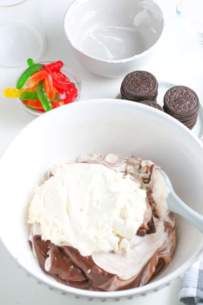 A mixing bowl with whipped topping being folded into chocolate pudding, with a spatula for chocolate dirt cups, next to Oreo cookie and gummy worms on the counter.