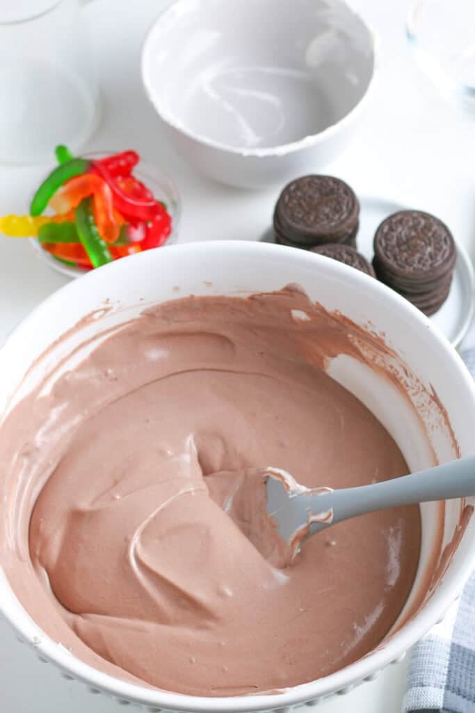 A bowl of chocolate puddding with cool whip after being mixed in for chocolate dirt cups next to sandwich cookies, gummy worms nearby.