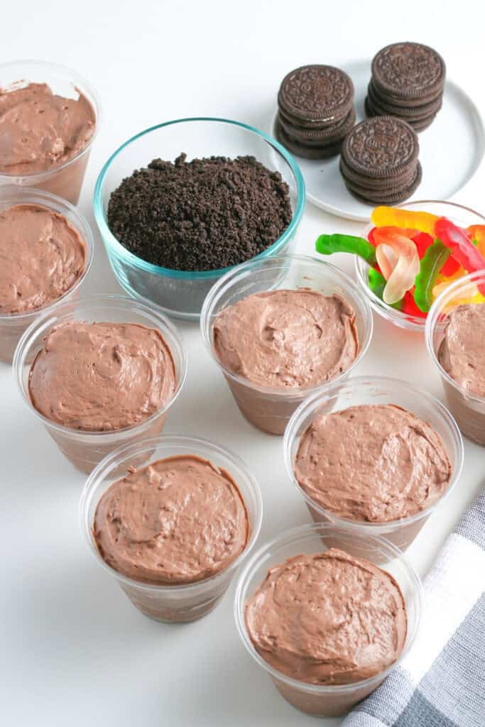 Eight plastic cups to make chocolate dirt cups filled with chocolate pudding, a bowl of crushed cookies nearby, whole sandwich cookies on a plate, and gummy worms.
