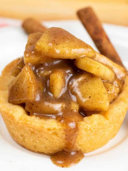 Close up side view of a sugar cookie cup filled with a gooey apple filling with some drizzling out on the plate below from the side next to a few sticks of cinnamon.